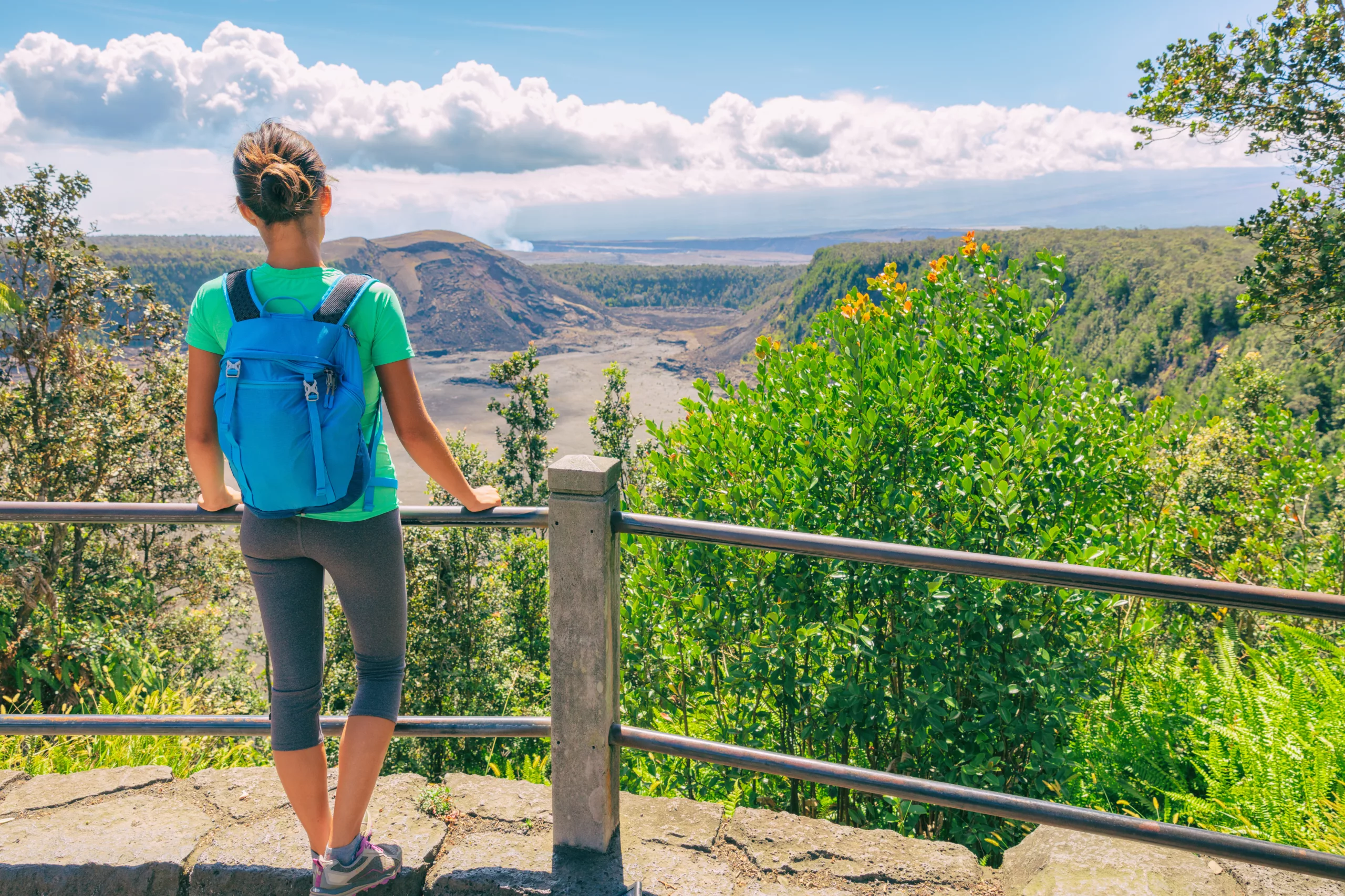 A female hiker wearing a backpack stands looking into Kilauea's caldera from a viewing platform on the Iki Trail