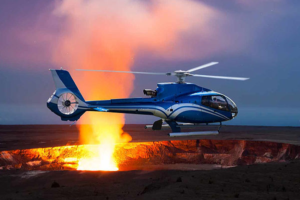 Blue helicopter flying over active volcano with orange lava erupting from it on a volcano tour from Oahu, Hawaii