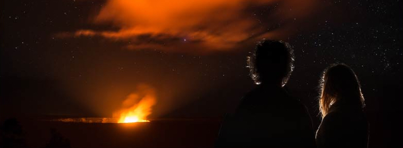 Silhouettes of a man and woman in front of active erupting volcano at night illuminated by bright orange lava